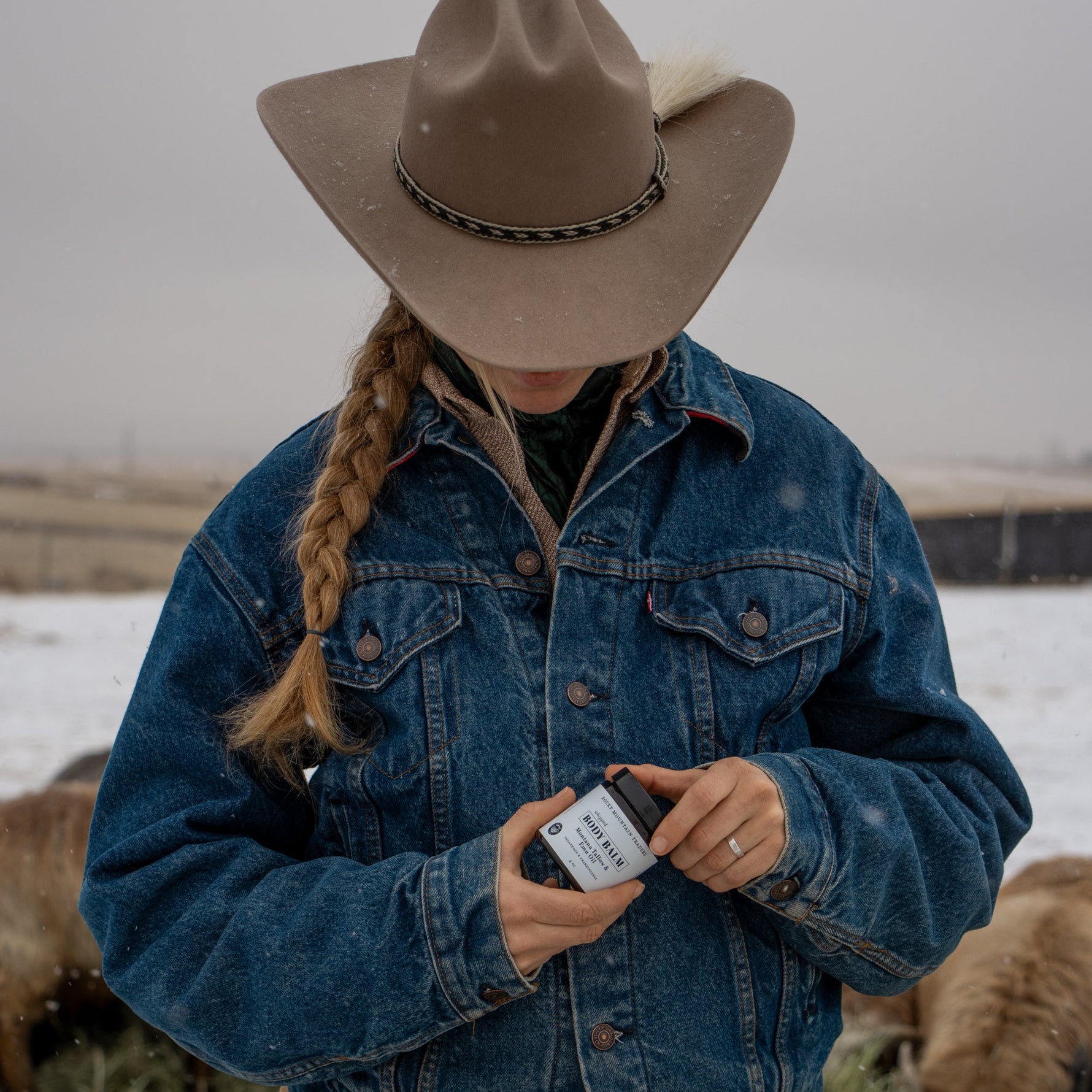 Person wearing a blue denim jacket and brown cowboy hat in a field with sheep, holding a jar of Body Balm.
