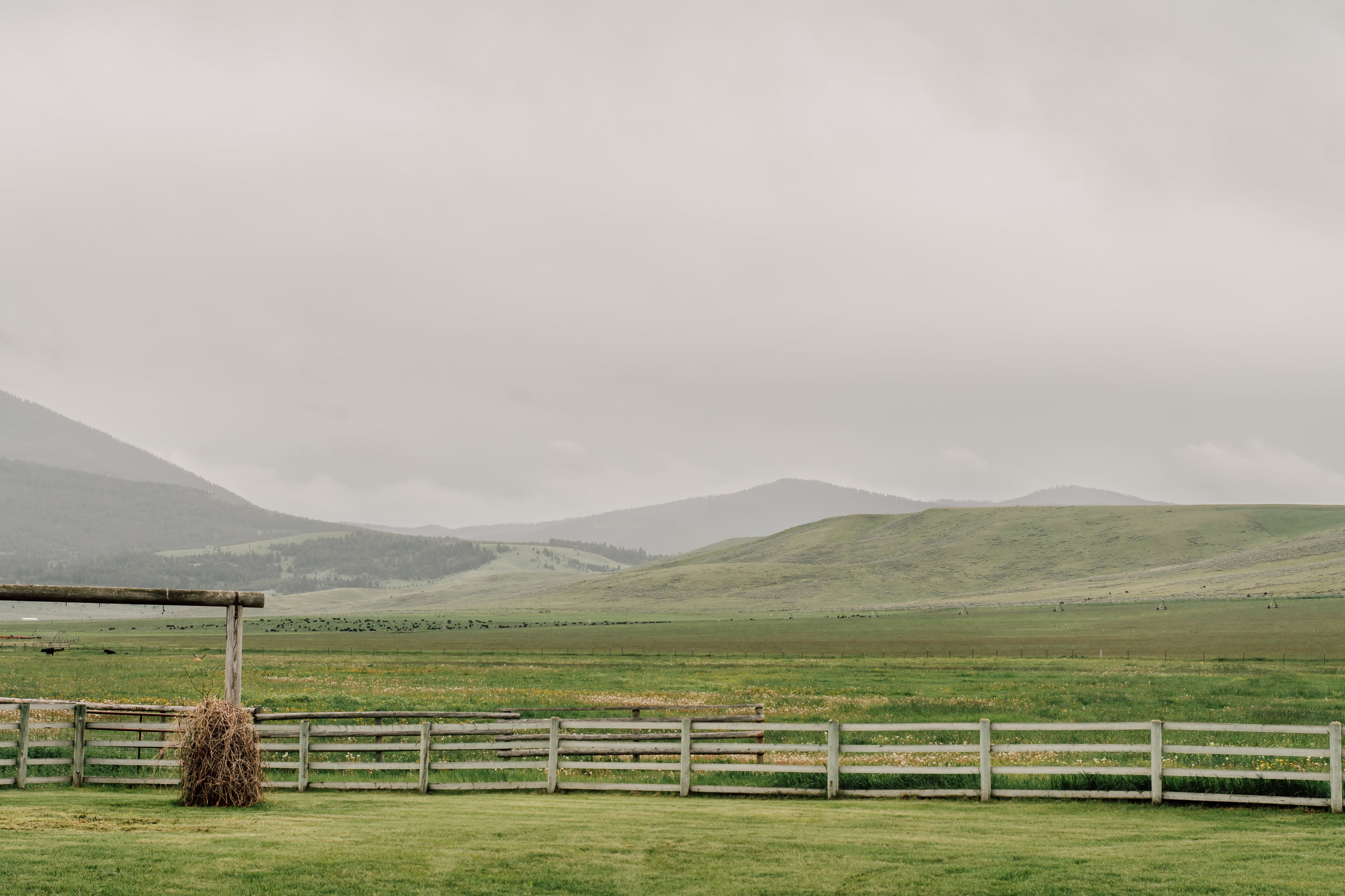 Rural landscape with a wooden fence, hay bale, and distant hills under an overcast sky.